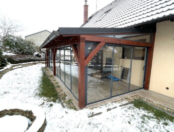 Alt text: Realizace rámového zasklení PLUS v barvě antracit RAL 7016 na dřevěné pergole u rodinného domu.