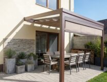 Detailní pohled na hnědou hliníkovou pergolu s baldachýnem a zataženou čelní screenovou roletou, která věrně napodobuje vzhled dřevěné pergoly 6X4.